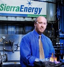 This image is a corporate photograph rather than a chart or diagram. It shows a man wearing glasses and a blue button-up shirt standing in an industrial facility, holding what appears to be a piece of
