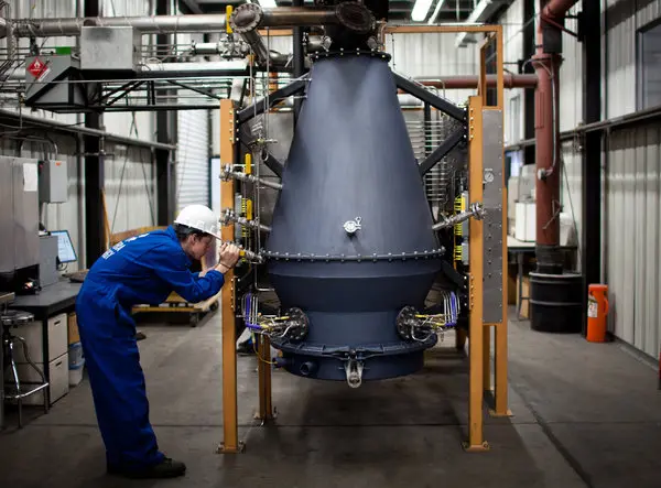 This photograph shows a technician wearing blue coveralls, a white hard hat, and safety glasses inspecting a large conical gasification vessel or reactor mounted on a yellow steel support frame within