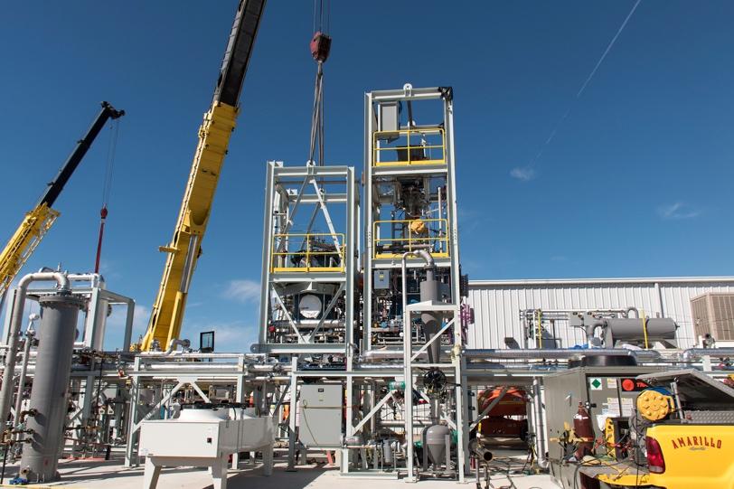 This image shows an industrial processing facility under construction, featuring a multi-level steel-framed structure with yellow safety railings and platforms. Two cranes—one yellow and one black—are