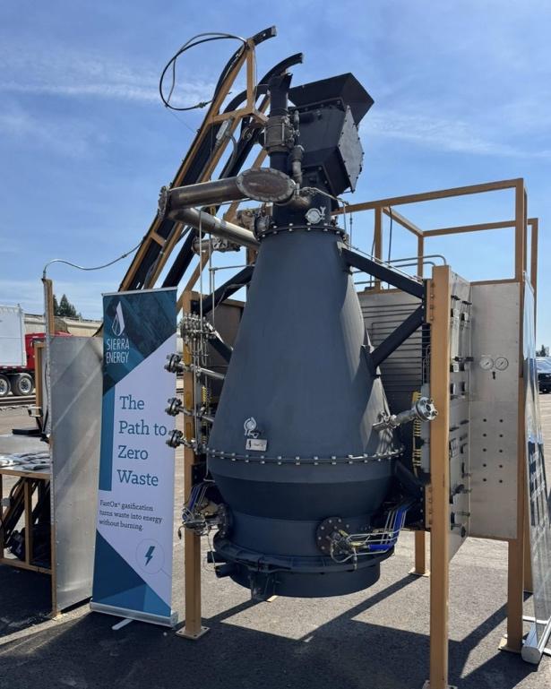 This image shows a Sierra Energy FastOx gasification unit—a large gray conical industrial reactor with numerous pipe connections, flanges, and instrumentation—displayed outdoors with a supporting yell